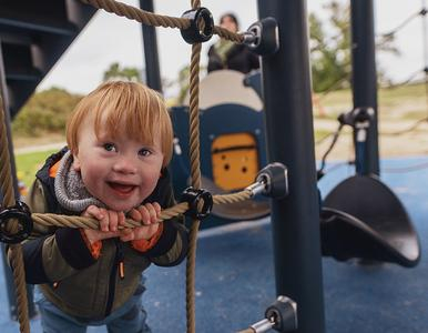 Ein Kind hält sich an den Seilen eines Klettergerüstes fest und lächelt in die Kamera. 