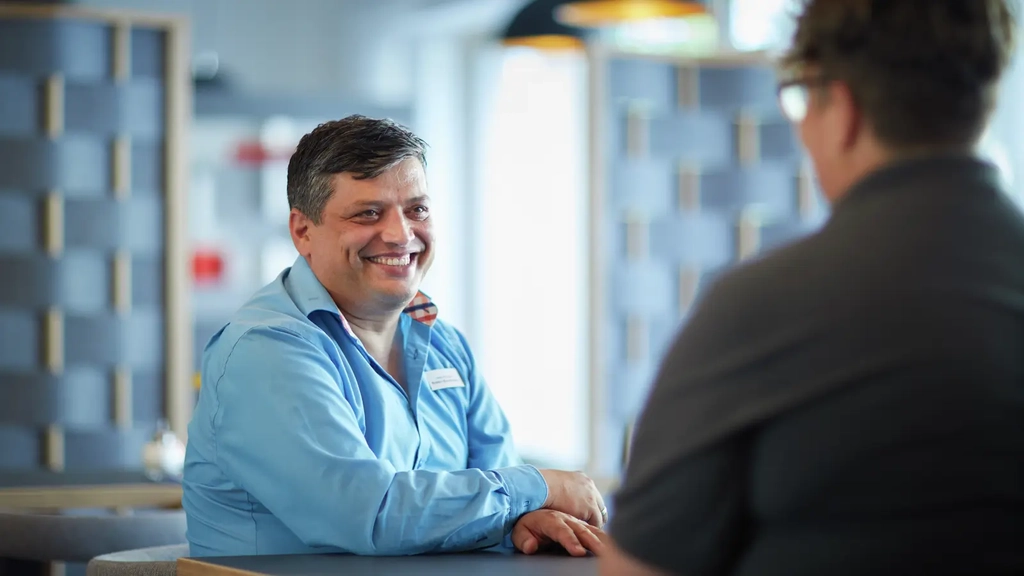 Ein Man im blauen Hemd unterhält sich mit einer dunkel gekleideten Person, die mit dem Rücken zur Kamera steht.