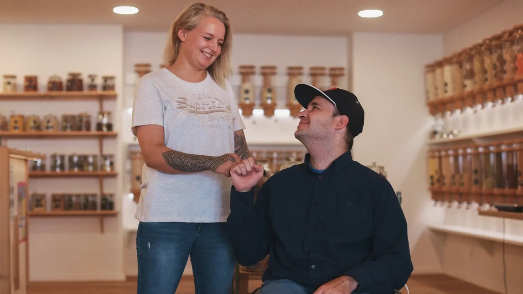 Eine Frau steht neben einem Mann im Rollstuhl, sie stoßen mit den Fäusten aneinander.