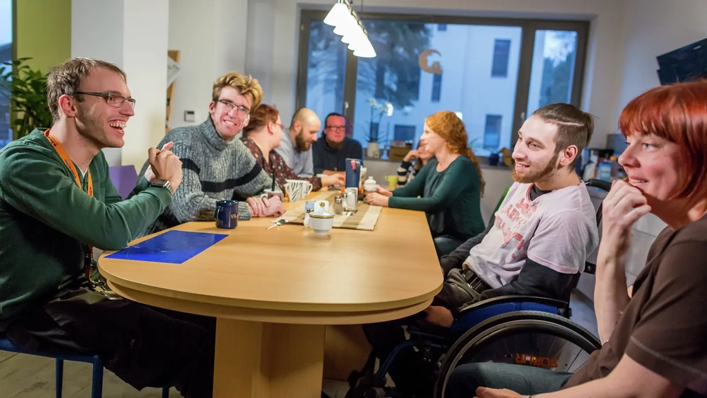 An einem Esstisch sitzen viele verschiedene Menschen mit und ohne Behinderung.