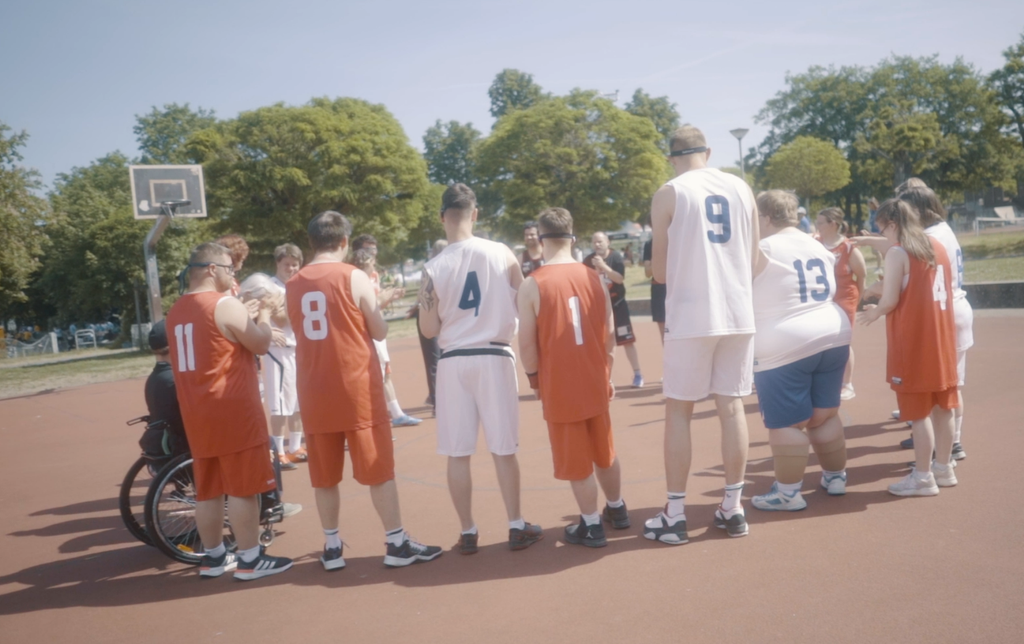 Eine Gruppe Basketballspieler mit und ohne sichtbare Behinderung stehen auf einem Basketballfeld im Kreis.