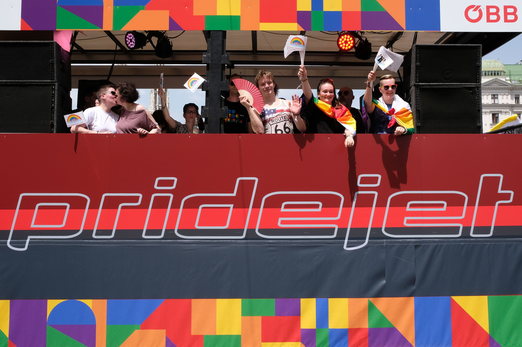 Junge Menschen mit Regenbogenfahnen stehen auf der Ladefläche eines Öbb-LKWs  mit der Aufschrift "pridejet" und winken.  Zwei Frauen küssen sich.