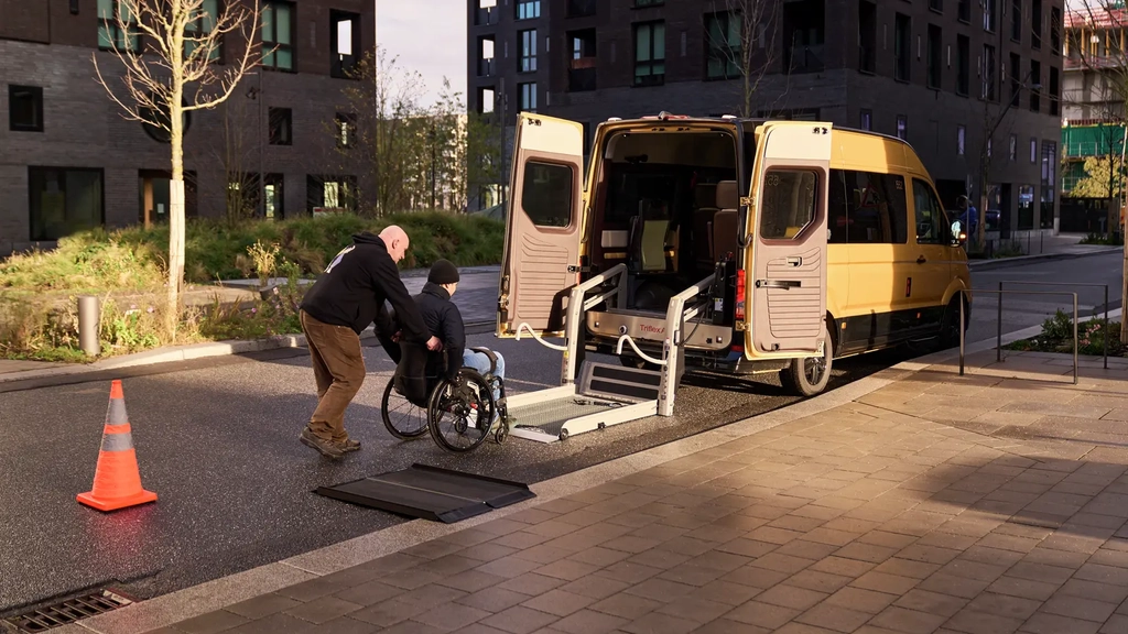 Ein MOIA Bus steht mit geöffneter Hecktür und heruntergelassener Rampe am Straßenrand. Ein Mann schiebt eine Person im Rollstuhl auf die Rampe.