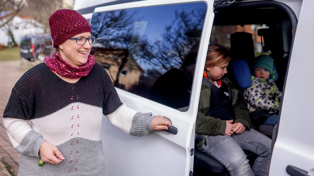 Eine Frau mit Wollmütze schließt die Schiebetür eines großen weißen Autos, in dem zwei Kinder sitzen.