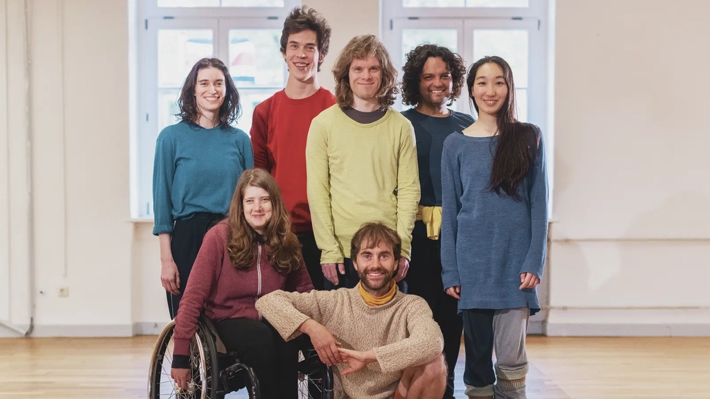 Eine Tanzkompanie aus Menschen mit und ohne Behinderung steht beieinander in einem Tanzsaal und lächelt in die Kamera.