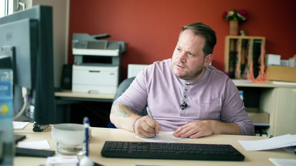 Ein Mann in einem lila T-Shirt sitzt an einem Schreibtisch im Büro. Er schaut auf den Computerbildschirm und schreibt etwas mit der Hand auf.