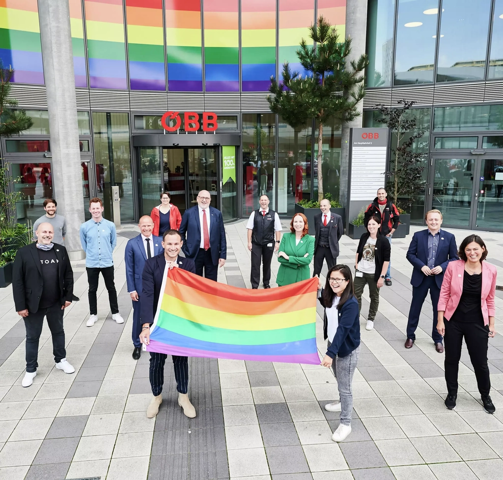 Frauen und Männer stehen vor dem ÖBB-Gebäude und lachen in die Kamera. Eine Frau und ein Mann halten eine Regenbogenfahne hoch.