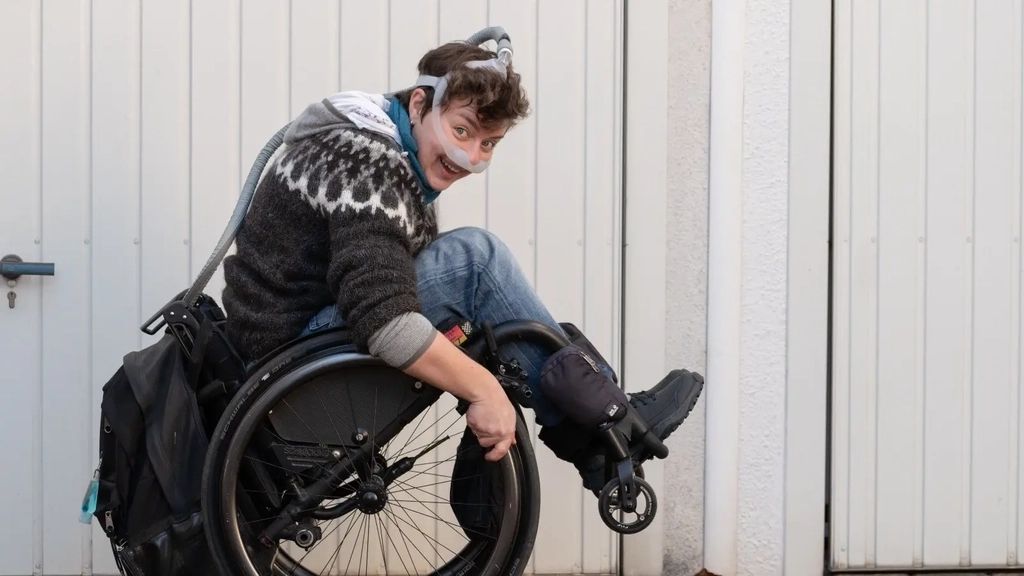 Eine junge Frau balanciert im Rollstuhl. Sie trägt ein Beatmungsgerät.
