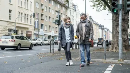 Ein Mann mit Langstock und eine Frau gehen über eine Straße.