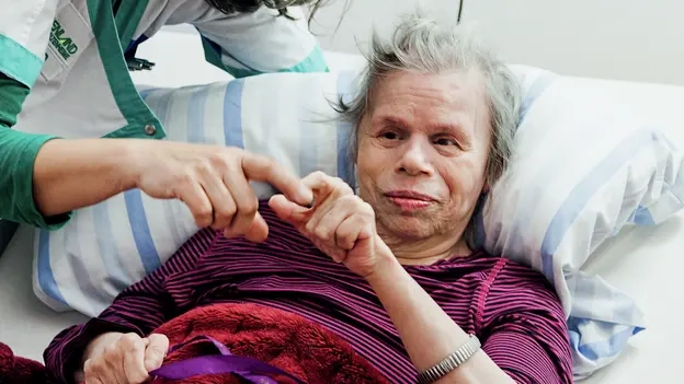 Rita Kohn liegt im Bett und macht eine Geste mit der Hand. Sabine Wuchold berührt mit ihrem Finger Ritas Finger. Sie versteht die Geste.