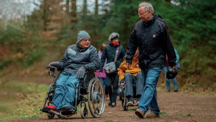 Zwei Männer sind im Wald unterwegs. Einer davon sitzt im Rollstuhl mit Joystick. Der andere spaziert daneben.