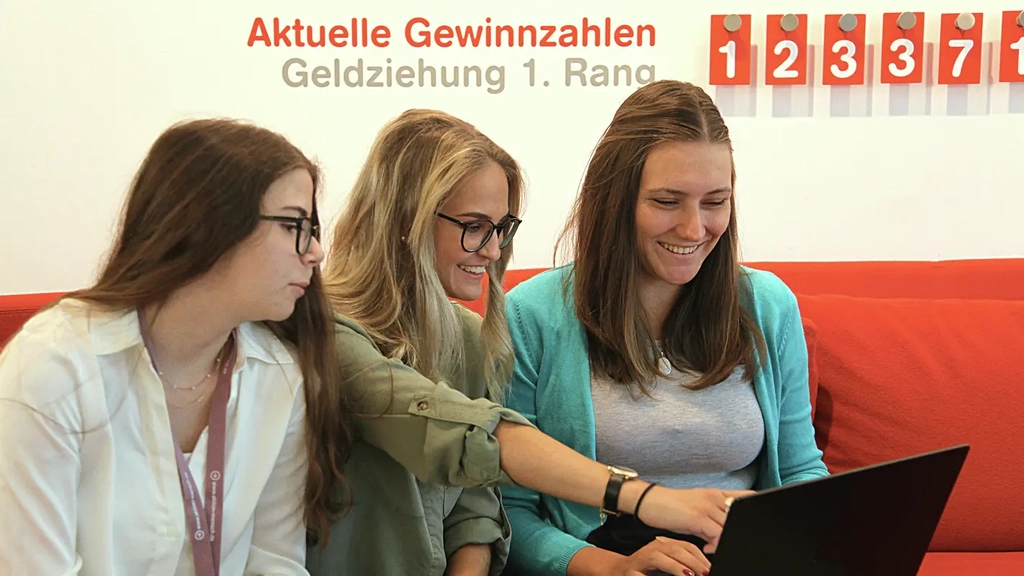 Drei junge Frauen sitzen auf einem roten Sofa und schauen gemeinsam auf einen Laptop. Hinter ihnen ist an der Wand der Text "Aktuelle Gewinnzahlen, Geldziehung 1. Rang" zu lesen. Daneben hängen weiße Ziffern auf roten Karten.