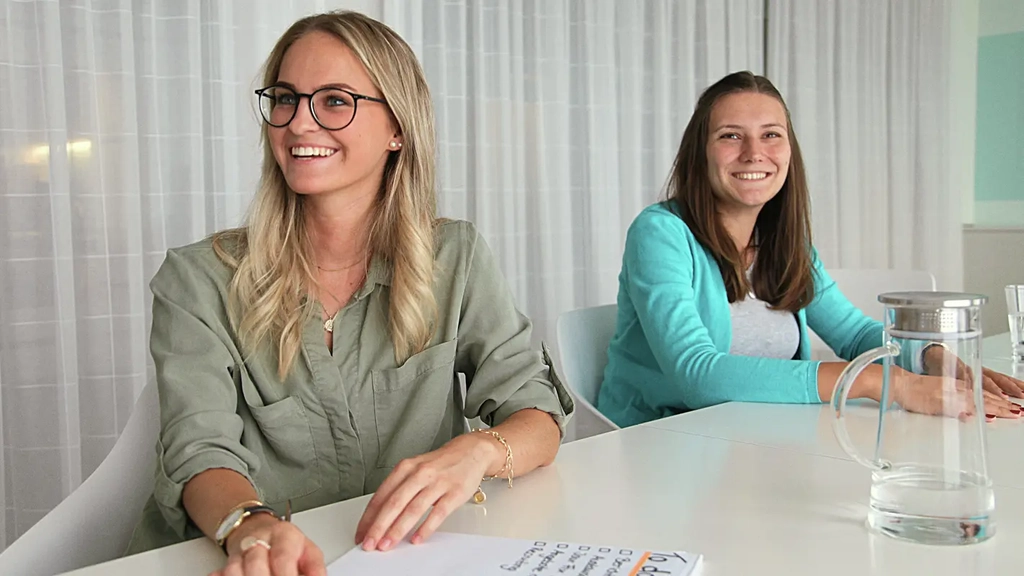 Zwei junge Frauen sitzen nebeneinander an einem großen Tisch in einem Konferenzraum. Die Vordere der beiden hat einen Block vor sich auf dem Tisch liegen. Dort ist die Überschrift "To Dos" zu lesen. Beide Frauen lachen freundlich.
