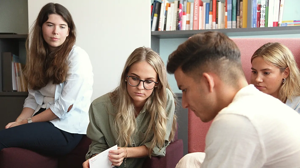 Drei junge Frauen und ein junger Mann sitzen gemeinsam in einer Sitzecke in einem Büroraum. Der junge Mann hält ein Smartphone in der Hand und zeigt den anderen darauf etwas. Die drei Frauen schauen aufmerksam hin.