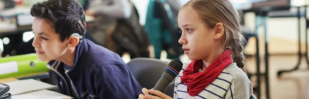 Ein Junge und ein Mädchen sitzen in einer Klasse. Das Mädchen spricht in ein Mikrofon. 