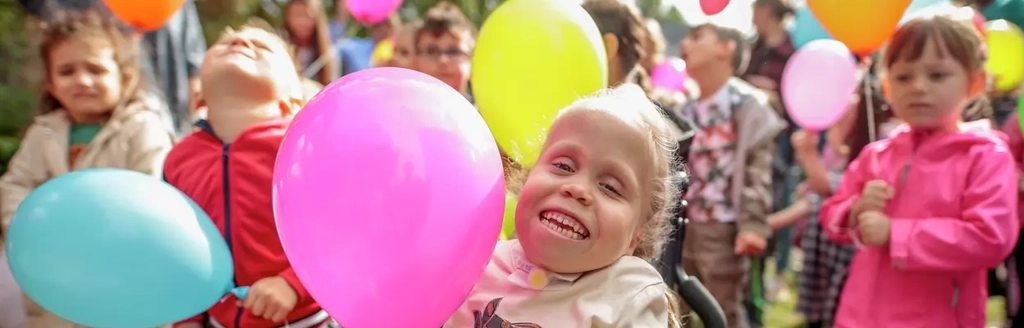 Mehrere Kinder stehen zusammen und halten Luftballons in den Händen. Ein Mädchen sitzt im Rollstuhl.