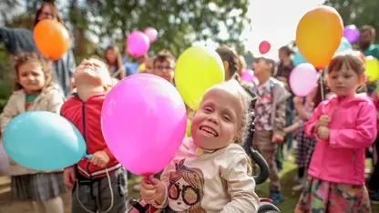 Mehrere Kinder stehen zusammen und halten Luftballons in den Händen. Ein Mädchen sitzt im Rollstuhl.
