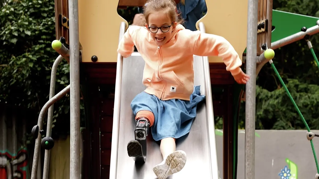 Ein Mädchen mit Beinprothese rutscht lächelnd eine Rutsche herunter.