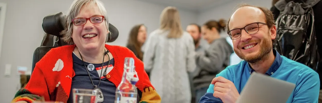 Eine Frau im Rollstuhl und ein Mann lachen in die Kamera.