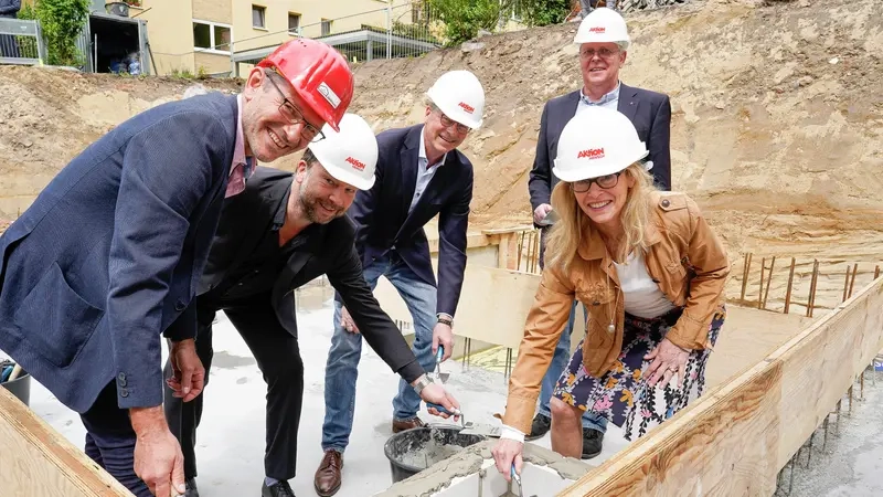 Eine Frau und vier Männer im Jackett und Bauhelm setzen einen Grundstein auf einer Baustelle