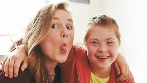 Zwei jugendliche Freundinnen mit und ohne Down-Syndrom Arm in Arm. Eine lacht in die Kamera, die zweite zieht eine lustige Grimasse.