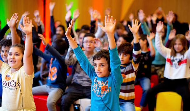 Mehrere Kinder sitzen auf bunten Hockern und strecken ihre Arme in die Höhe.