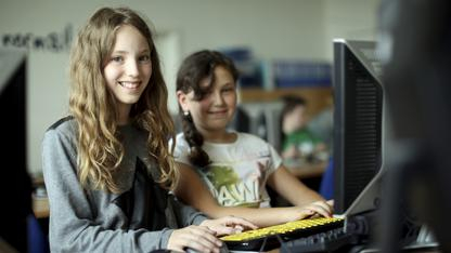 Zwei Mädchen sitzen vor einem Rechner. Beide lächeln in die Kamera.