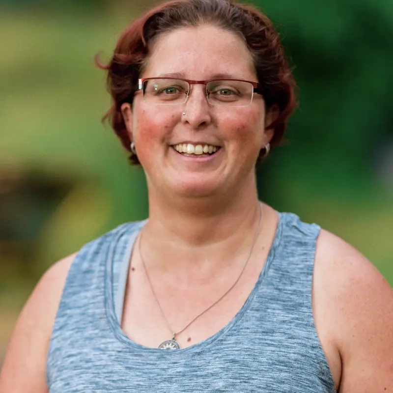 Eine Frau im grauen Tanktop steht in einem Garten. Sie trägt eine Brille mit schmalem Rahmen, eine Smartwatch und hat kurzes rötliches Haar. Sie schaut in die Kamera und lächelt entspannt.