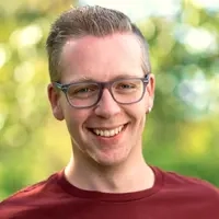 Ein junger Mann steht in einem Garten. Er hat kurzes, blondes Haar und trägt eine Brille. Er schaut in die Kamera und lächelt freundlich.