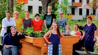 Eine Gruppe von Menschen mit und ohne Behinderung steht um ein Hochbeet, das sie gerade bepflanzt haben. Alle lächeln.