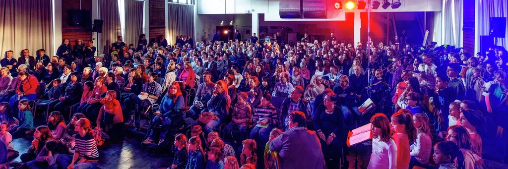 Viele Kinder und Jugendliche sitzen in einer großen, verdunkelten Halle in Reihen und schauen nach vorne. Am Ende des Saals ist ein Scheinwerfer angebracht, der rotes Licht  über die Zuschauer strahlt.