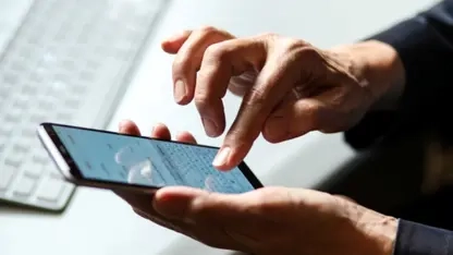 zwei haende bedienen ein smartphone. im hintergrund ist ein schreibtisch mit tastatur und computermaus zu sehen.