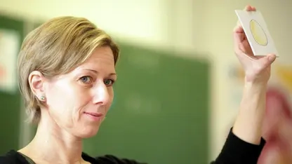 Eine Frau mit kurzen blonden Haaren hält eine Karte mit einem gezeichneten Ei in die Höhe. Im Hintergrund sieht man unscharf eine Schiefer-Tafel.