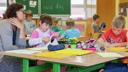Lehrerin und drei Kinder an einem Tisch in einem Klassenzimmer. Im Hintergrund sieht man eine Tafel.