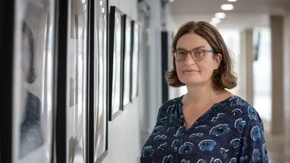 Eine Frau mit braunen Haaren und Brille steht vor einer Bildergalerie im Flur einer Schule.