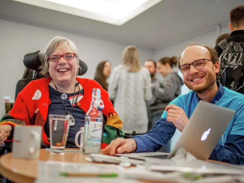 Eine Frau im Rollstuhl und ein Mann lachen in die Kamera.