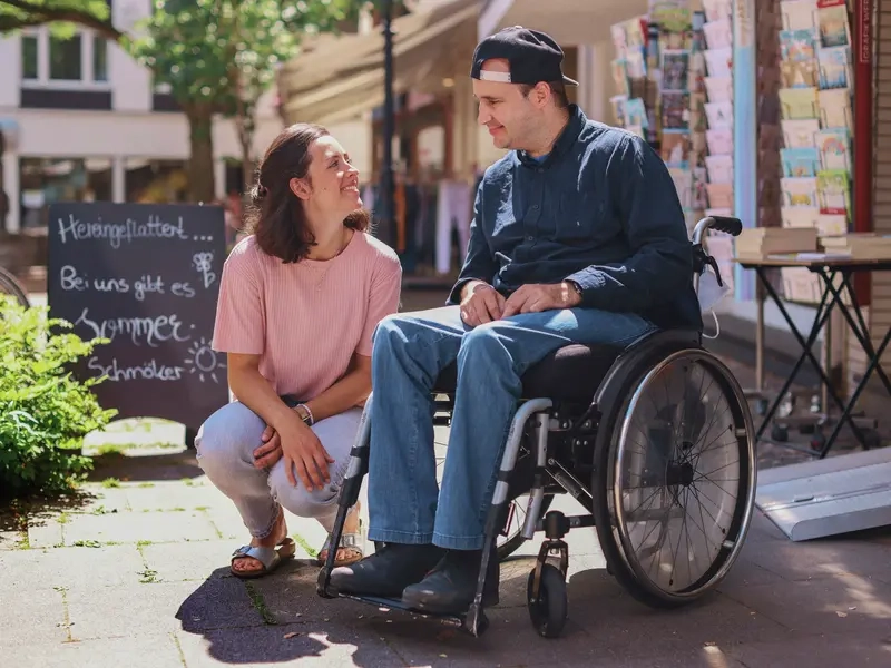 Ein Mann im Rollstuhl sitzt vor einer Buchhandlung. Neben ihm sitzt eine Frau in der Hocke. Die beiden lächeln sich an.