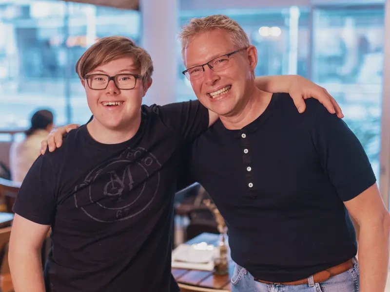 Ein Mann mit Down-Syndrom und ein Mann mit Brille haben einen Arm um die Schultern des anderen gelegt.