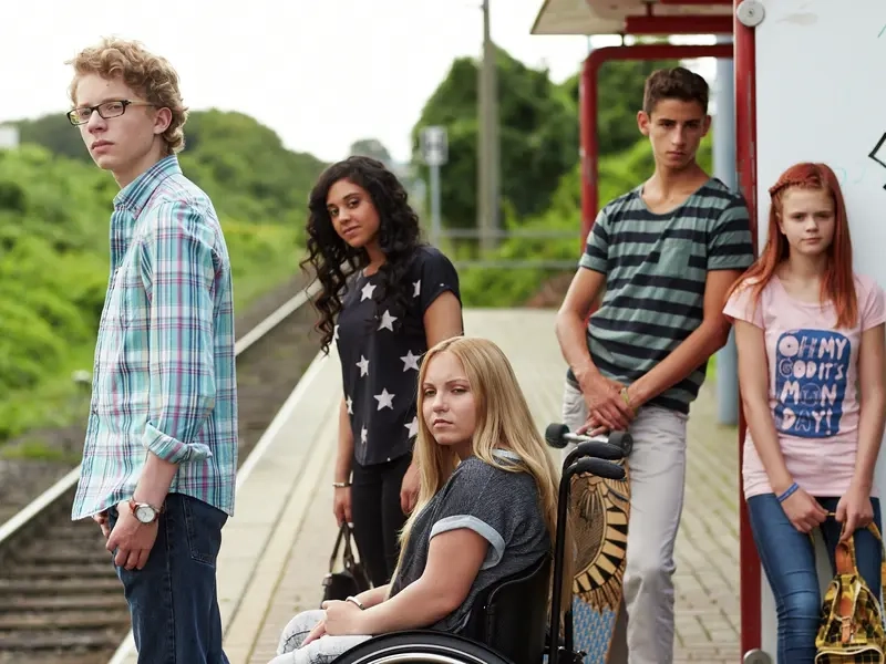 Fünf Jugendliche, zwei Jungen und drei Mädchen, stehen an einem Bahnsteig. Ein Mädchen sitzt im Rollstuhl.