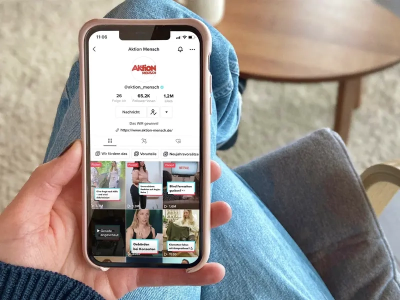 Blick von oben auf ein Handy in der Hand einer Frau. Auf dem Display ist der TikTok-Account der Aktion Mensch zu sehen.