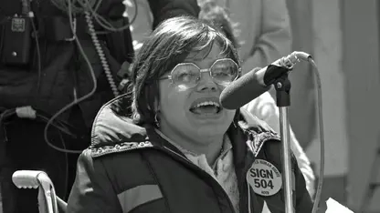 Das historische schwarz-weiße Bild zeigt eine Frau im Rollstuhl, die bei einer Kundgebung ins Mikrofon spricht.