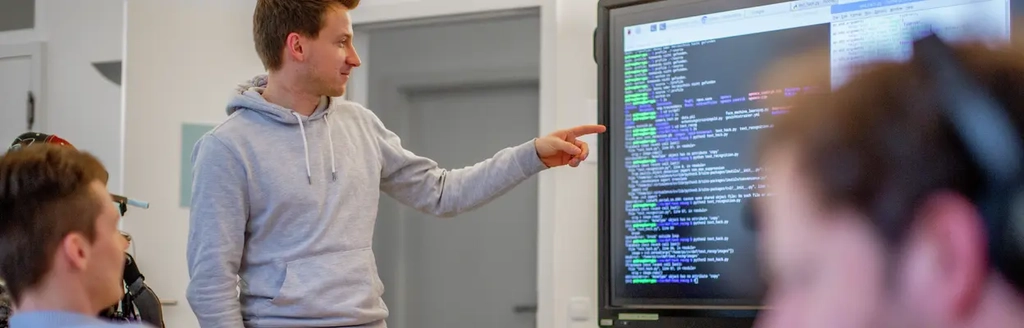 Ein junger Mann zeigt auf einen Bildschirm auf dem Programmiercode zu sehen ist. Ein weiterer junger Mann schaut ebenfalls zu diesem Bildschirm.