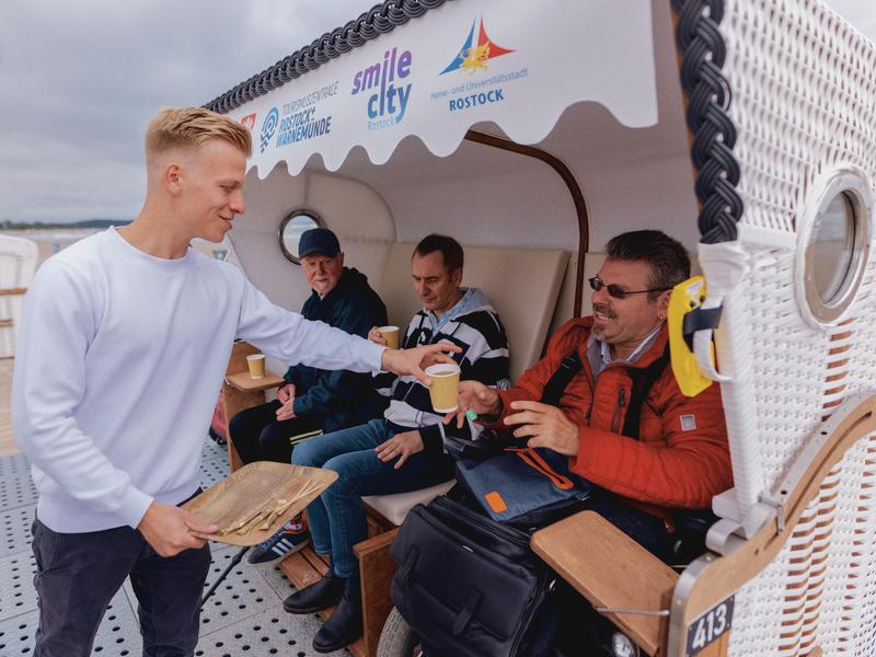 Drei Männer sitzen in einem Strandkorb. Eine Person sitzt im Rollstuhl. Sie bekommen einen Kaffee serviert.