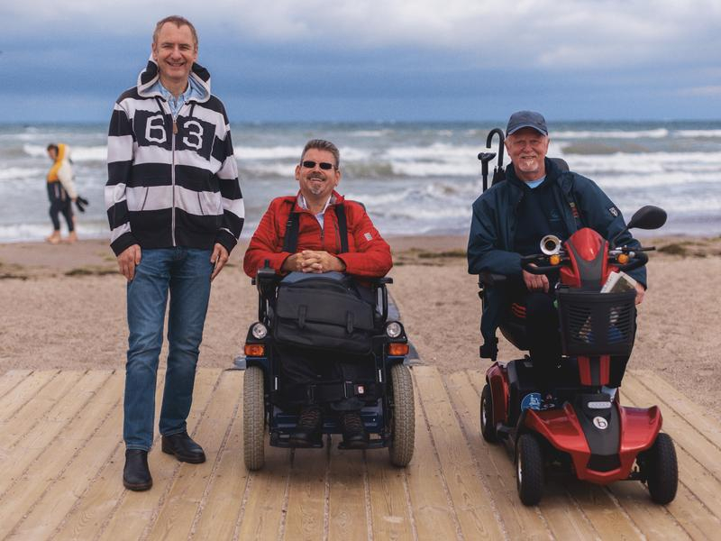 Drei Männer, zwei davon im Rollstuhl, stehen auf einem Steg am Strand und blicken aufs Meer.