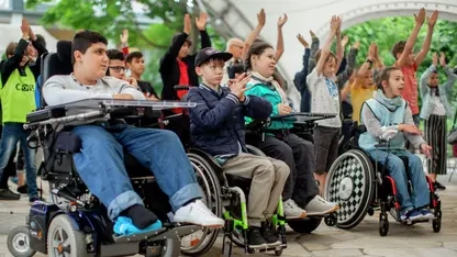 Mehrere Kinder sitzen und  stehen in mehreren Reihen. Manche davon sitzen im Rollstuhl. Die meisten von ihnen jubeln.