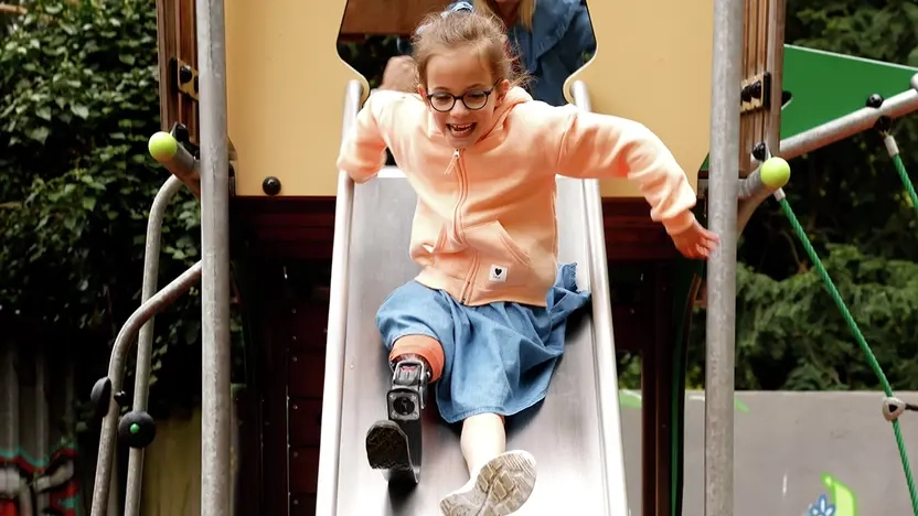 Ein Mädchen mit Beinprothese rutscht lächelnd eine Rutsche herunter.