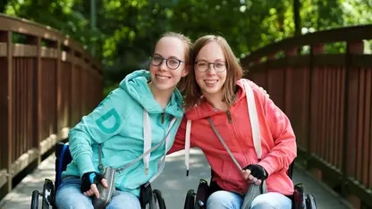 Leona und Ellen lachen in die Kamera. Sie sitzen beide Rollstuhl.