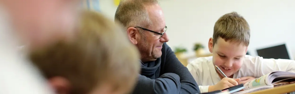 Ein Junge schreibt etwas in ein Schulheft. Neben ihm sitzt ein Erwachsener und schaut ihm dabei zu. Im Vordergrund sind unscharf zwei weitere Kinder zu sehen.