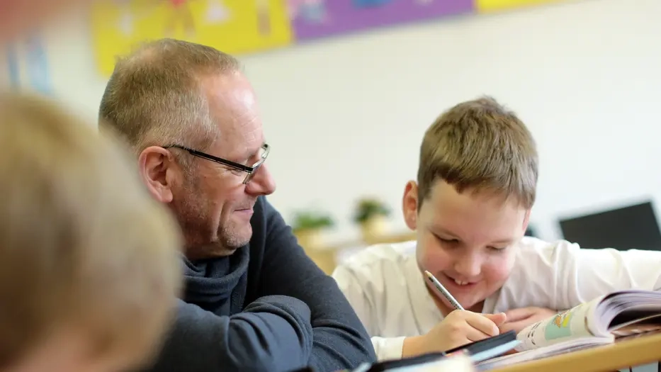 Ein Junge schreibt etwas in ein Schulheft. Neben ihm sitzt ein Erwachsener und schaut ihm dabei zu. Im Vordergrund sind unscharf zwei weitere Kinder zu sehen.