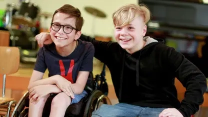 Zwei Jungen sitzen nebeneinander. Der eine auf einem Stuhl, der andere in einem Rollstuhl. Der Junge auf dem Stuhl hat den Arm um den Jungen im Rollstuhl gelegt. Beide lachen in die Kamera.